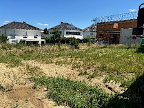 Blick auf ein unbebautes Grundst&uuml;ck mit hohem Gras und umliegenden H&auml;usern unter blauem Himmel.